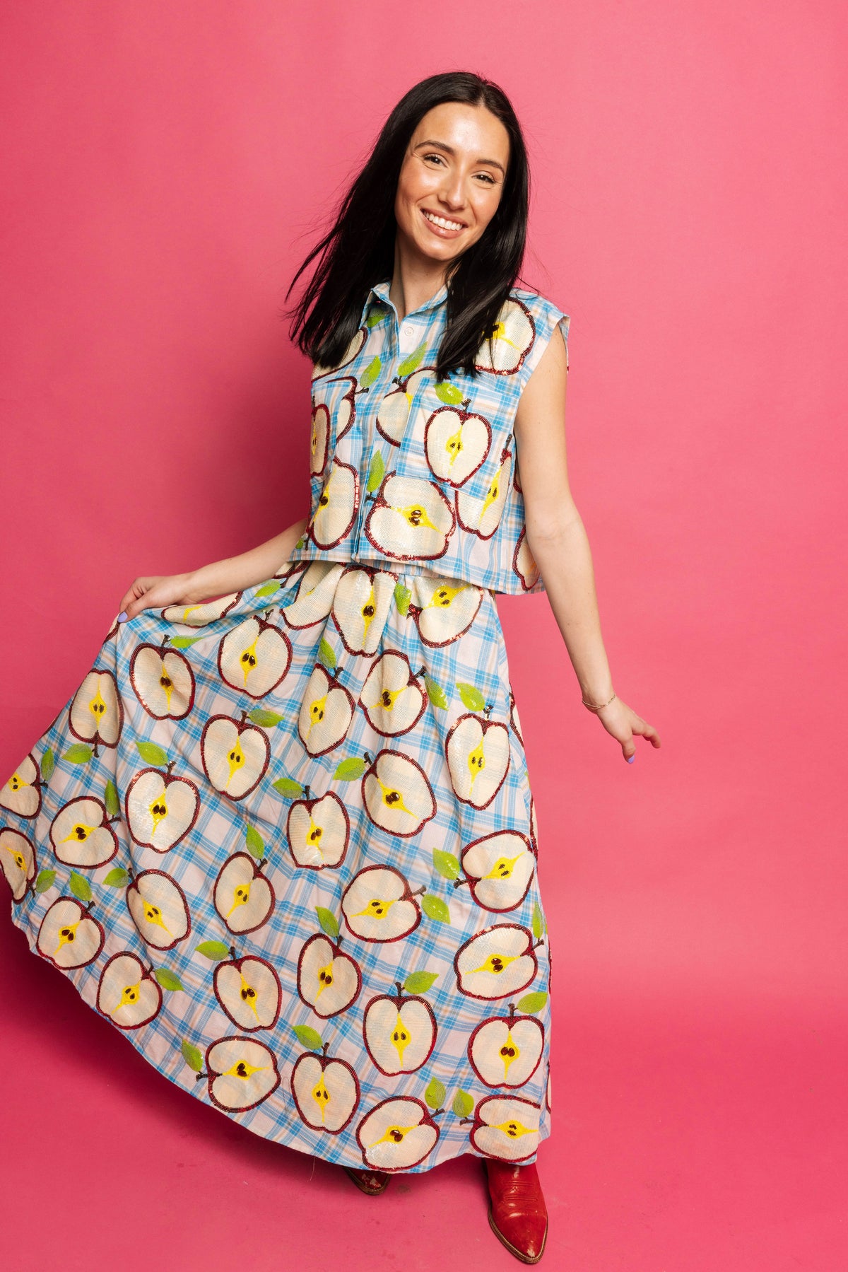 A smiling model holds out the matching blue plaid apple-slice maxi skirt while wearing the Queen of Sparkles sleeveless button-up tank against a pink backdrop.