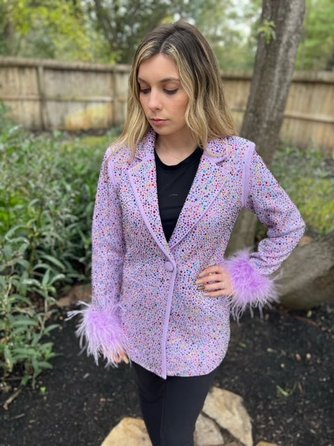 Model wearing a lavender Queen of Sparkles rhinestone blazer with feather cuffs and a black top outdoors.