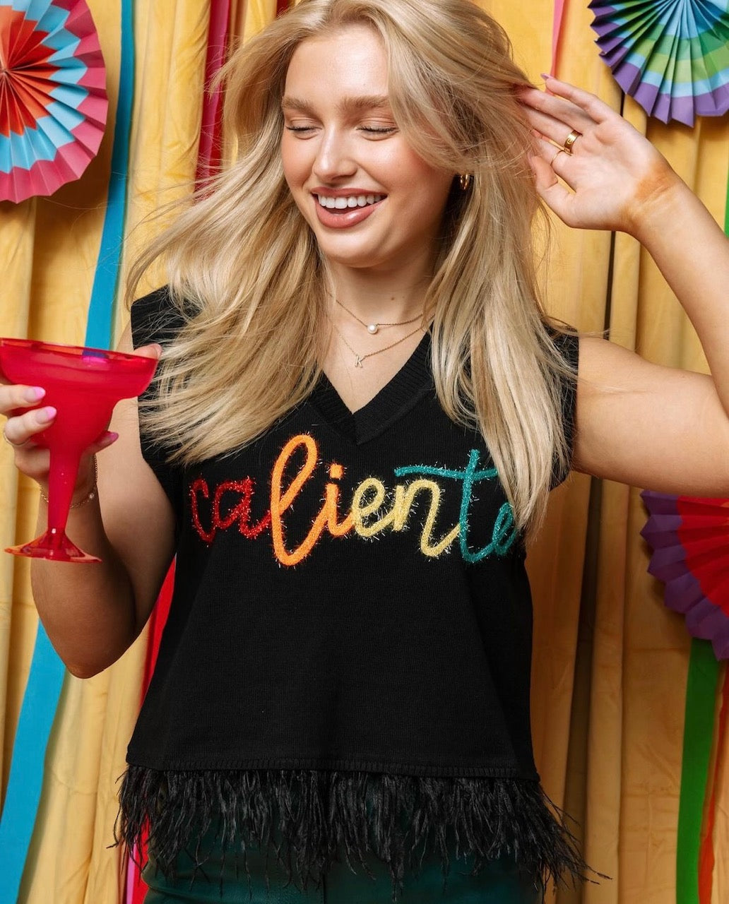 Close-up of a model wearing the black Caliente tank with colorful lettering and a feathered hem, holding a red cup.