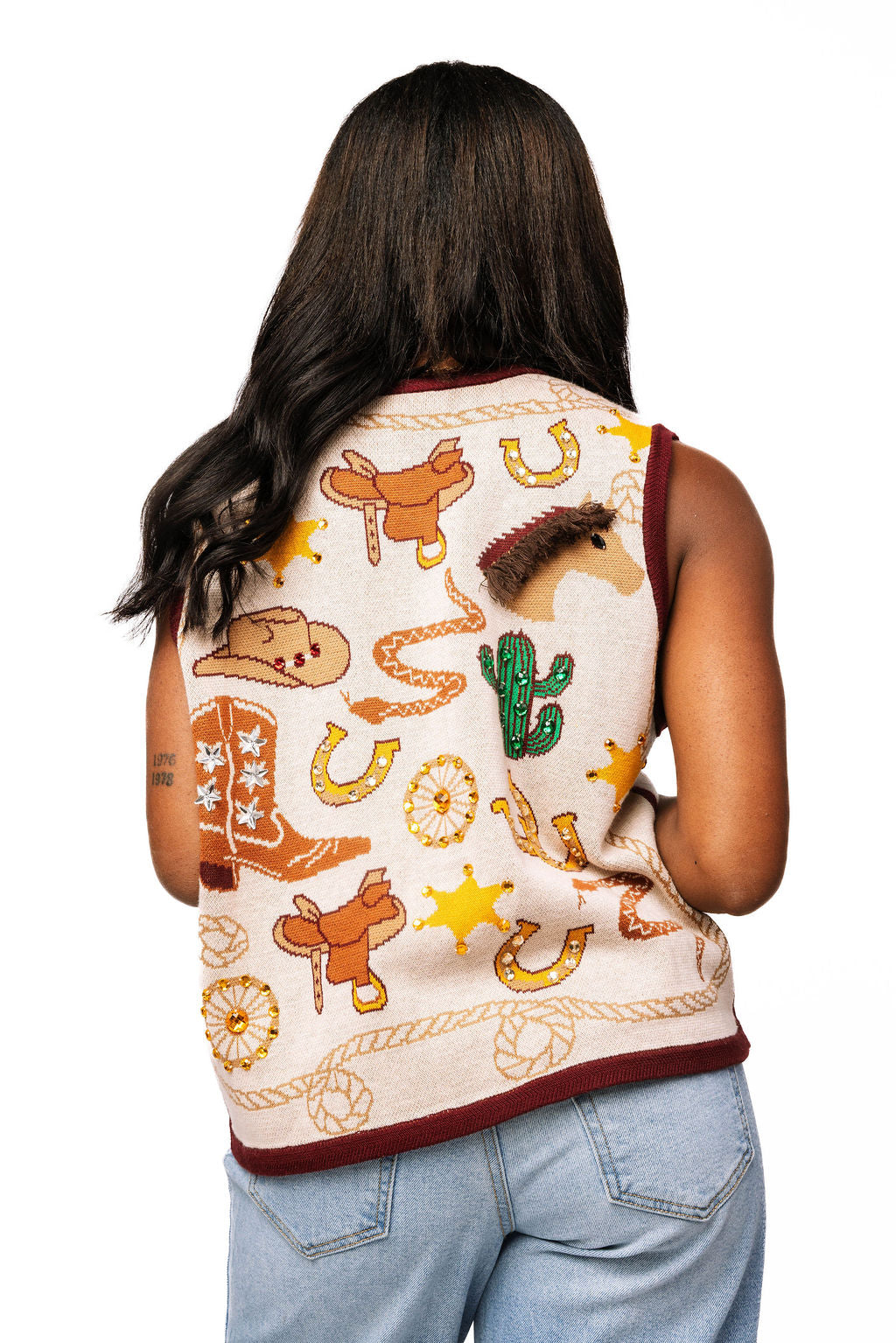 Back view of a model wearing an ivory knit vest with burgundy trim and Western icons, including cowboy boots, horseshoes, and a cactus.