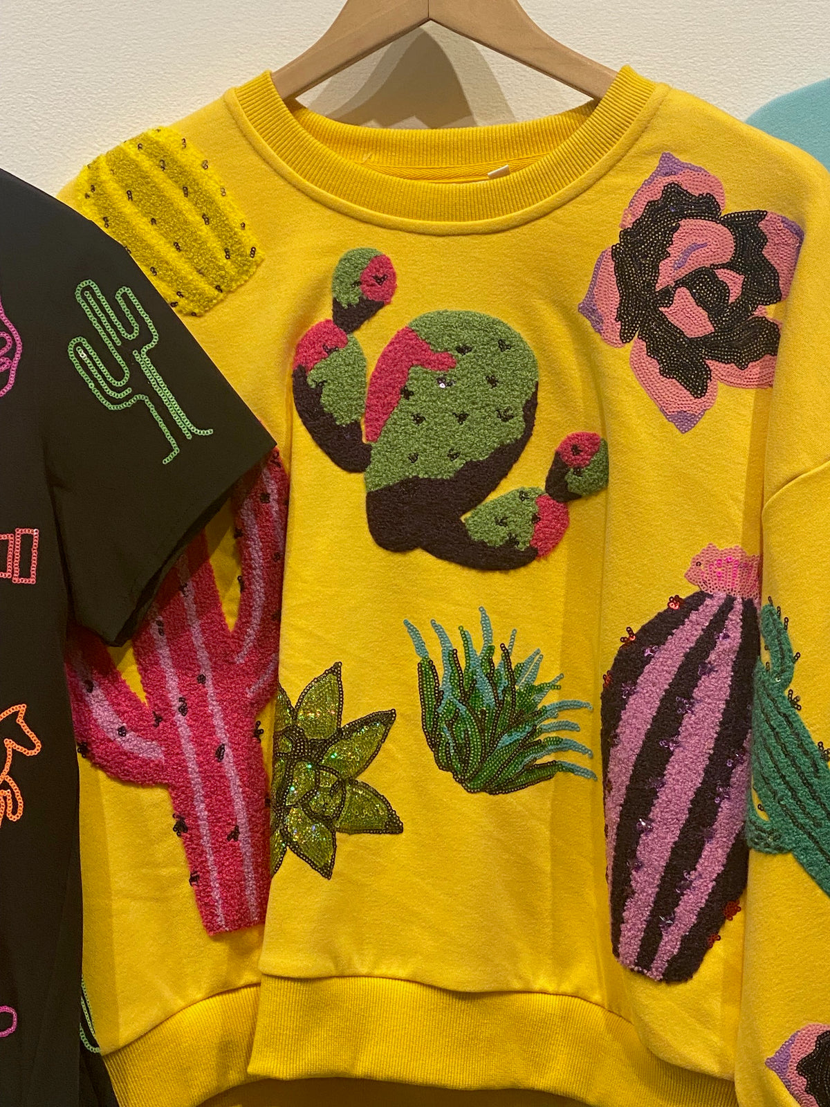 Close-up of the bright yellow Queen of Sparkles sweatshirt on a hanger, showing colorful cactus and desert-flower motifs with beaded-looking embellishments; other garments are partly visible beside it.