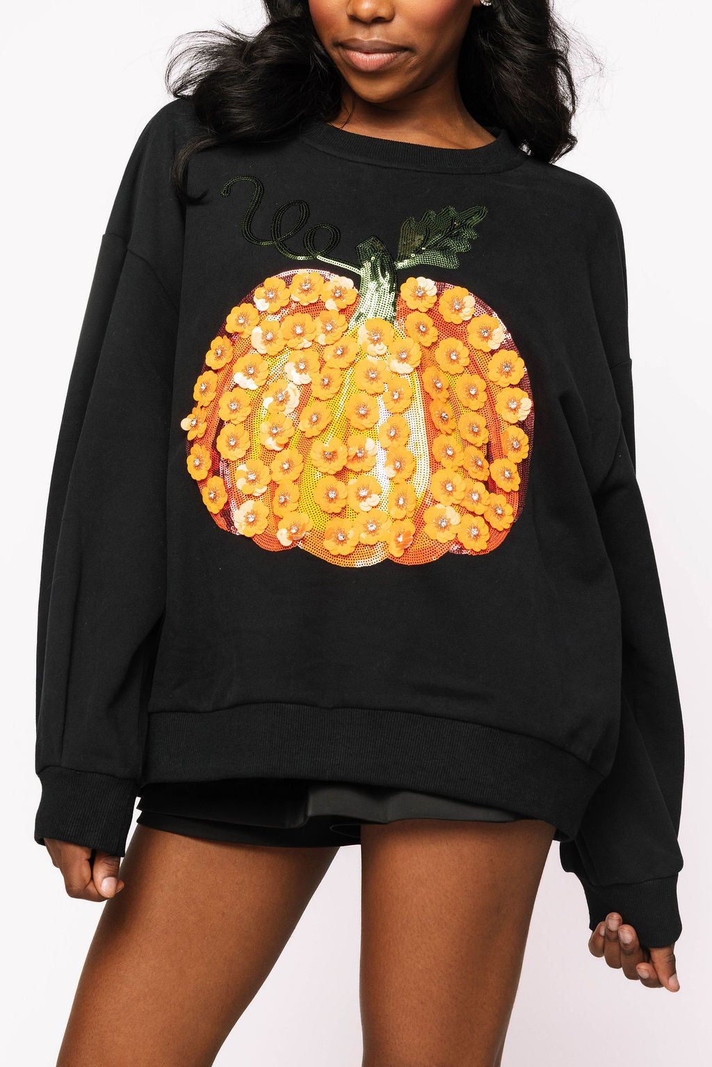 Front view of a model wearing the Queen of Sparkles black sweatshirt with an oversized orange pumpkin decorated with sparkling flower motifs and a green stem.