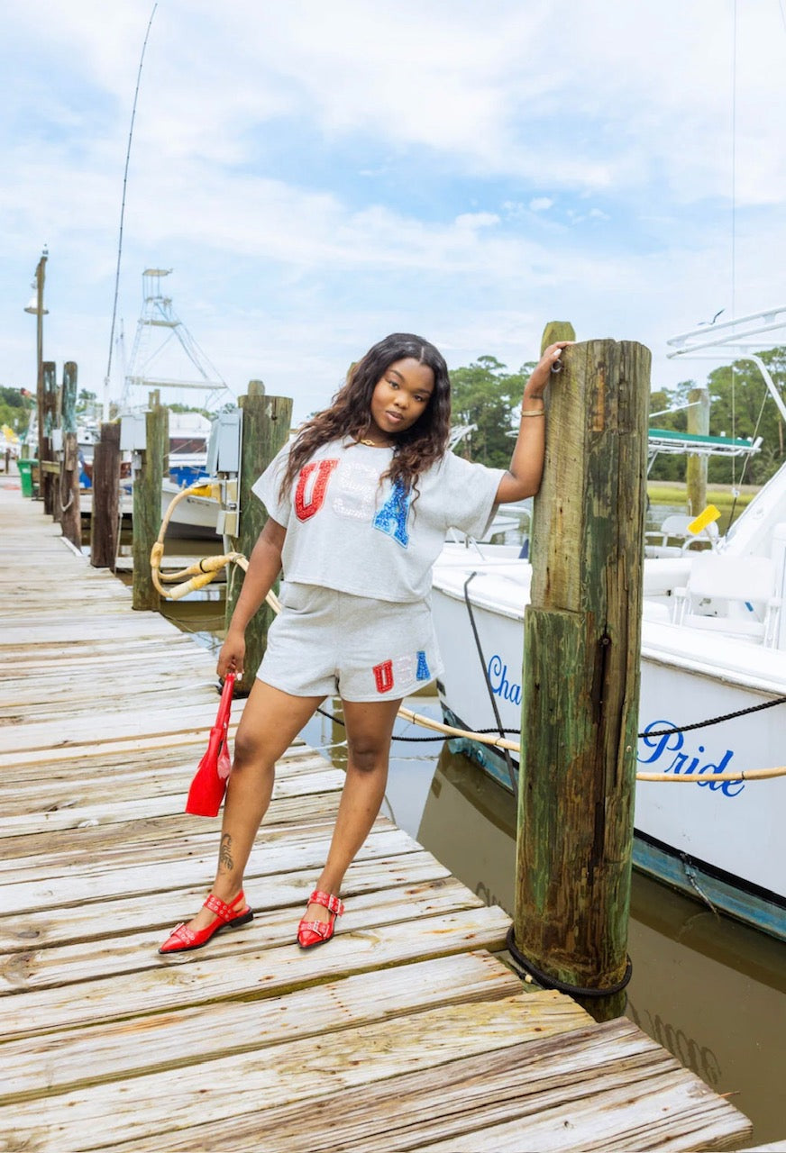 Model wearing grey USA embellished top with matching shorts and red accessories on a dock