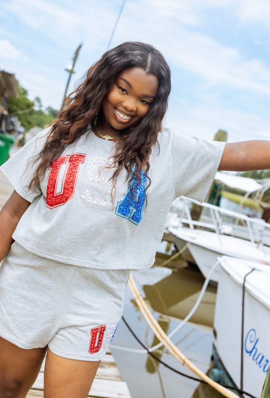 Model wearing grey cropped Queen of Sparkles USA top and matching shorts beside boats at a marina