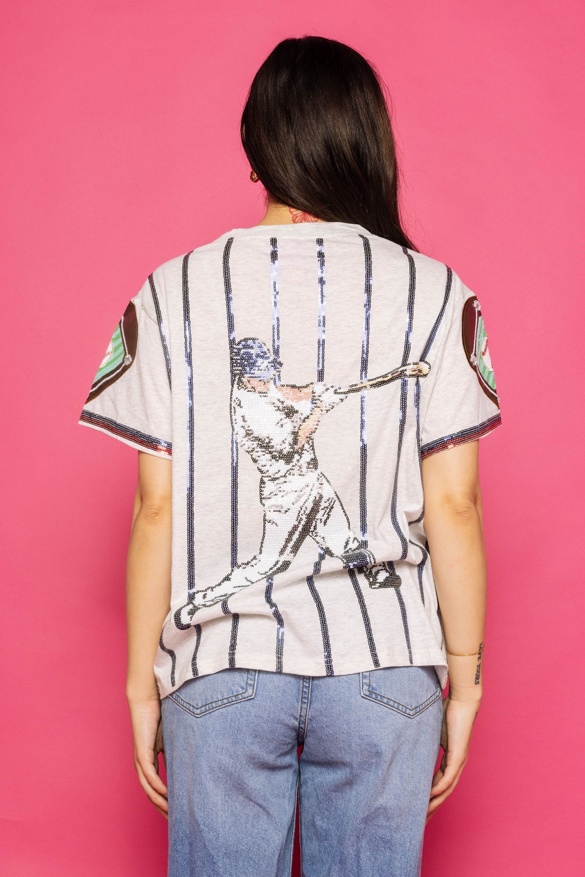 Back view of a model wearing the Queen of Sparkles grey and royal strike out baseball tee with vertical pinstripes, baseball-themed back graphic, and multicolor sleeve trim against a pink background.
