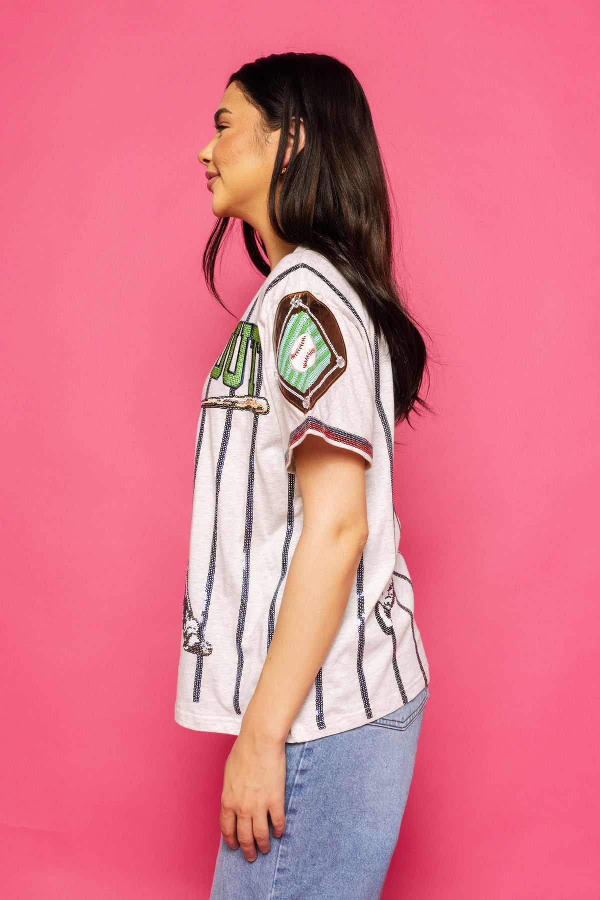 Side-profile view of a model wearing the Queen of Sparkles grey and royal strike out baseball tee with vertical stripes and a baseball-themed sleeve patch against a pink background.