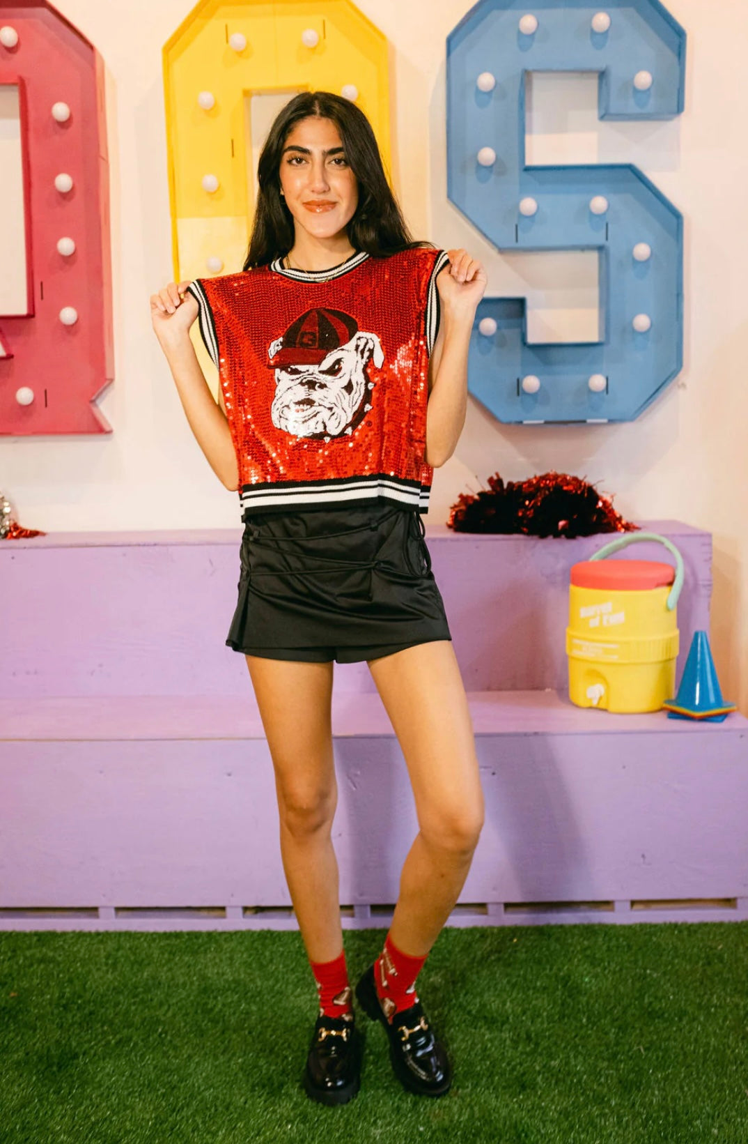 Model holding the hem of a red sequin bulldog-head sweater tank to show its shape and striped trim, with a black skirt.