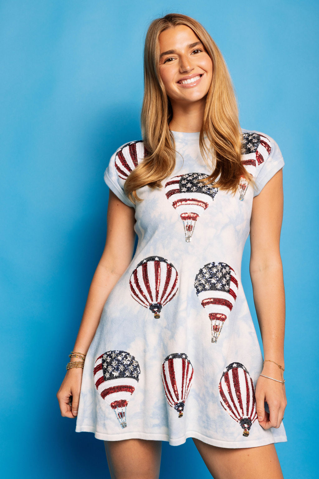 Model wearing the Queen of Sparkles light blue tie-dye Hot Air Balloon Sweater Dress, front view against a blue background, showing its short sleeves and embellished red, white, and blue balloon motifs.