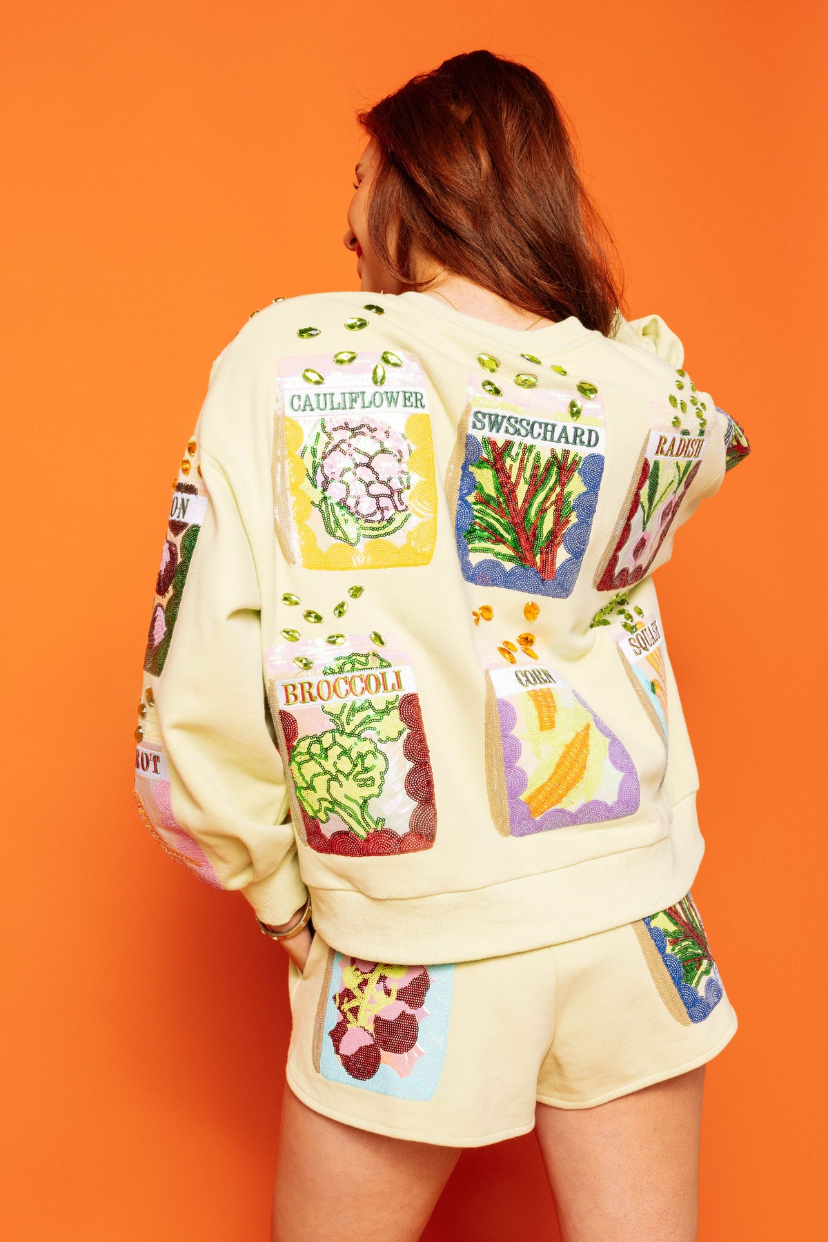 Back view of a model wearing the Queen of Sparkles cream V-neck sweatshirt with colorful seed-packet designs and embellished seed details on the back, styled with matching shorts against an orange background.