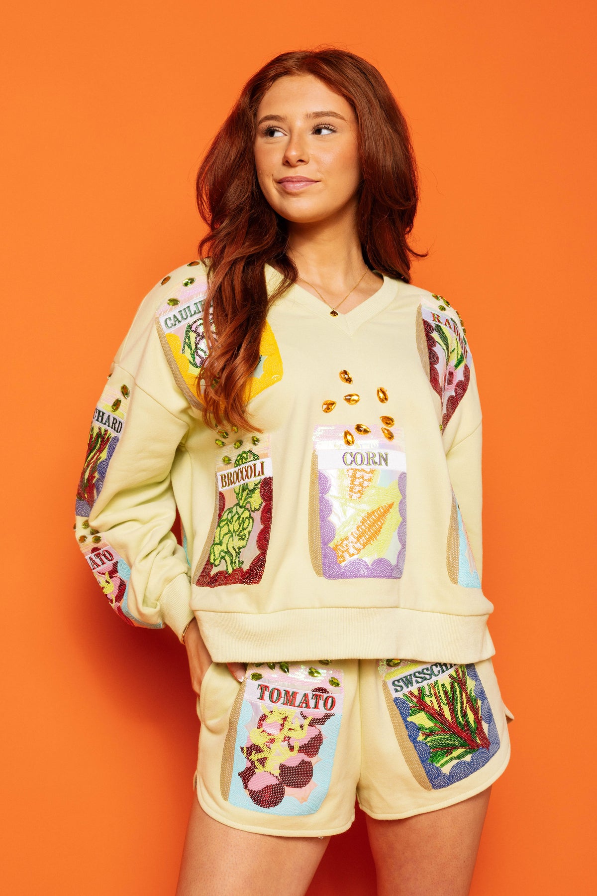 Model wearing the Queen of Sparkles cream V-neck sweatshirt with colorful seed-packet designs and embellished seed details on the front and sleeves, styled with matching shorts against an orange background.