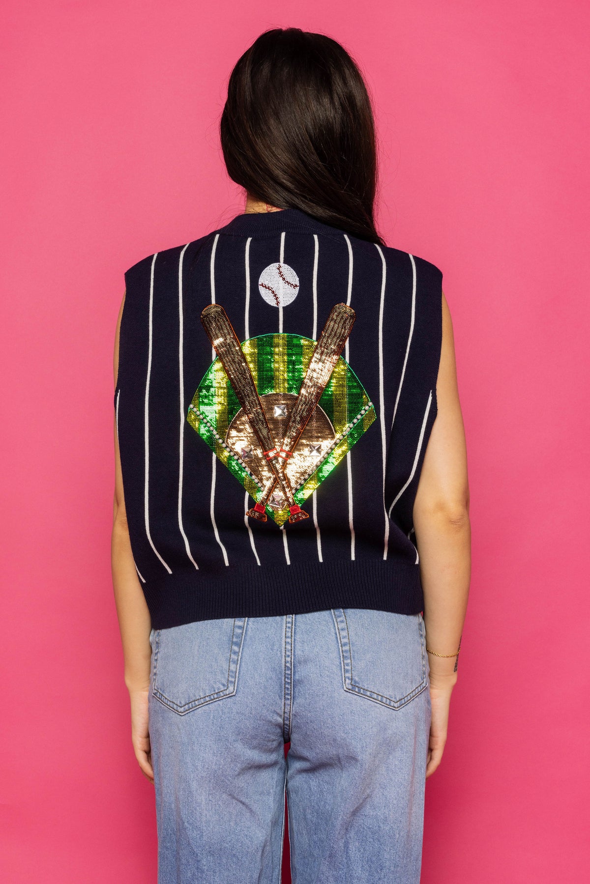 Back view of a model wearing the navy-and-white pinstriped Queen of Sparkles Batter Up sweater vest, showing its colorful embellished baseball design, against a pink background.