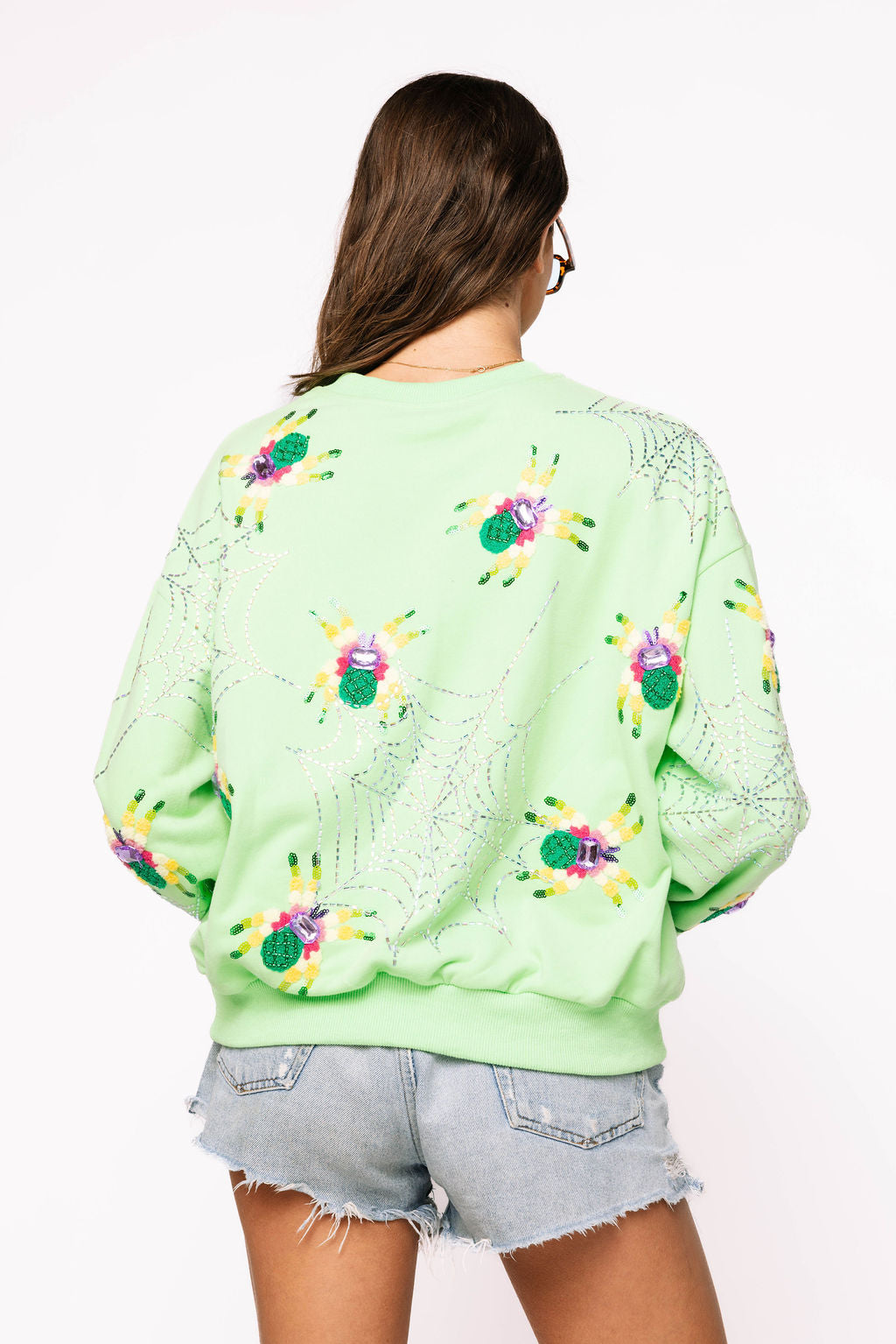 Back view of a model wearing the neon light green Queen of Sparkles sweatshirt, showing colorful fuzzy spider motifs and rhinestone spiderwebs across the back and sleeves.