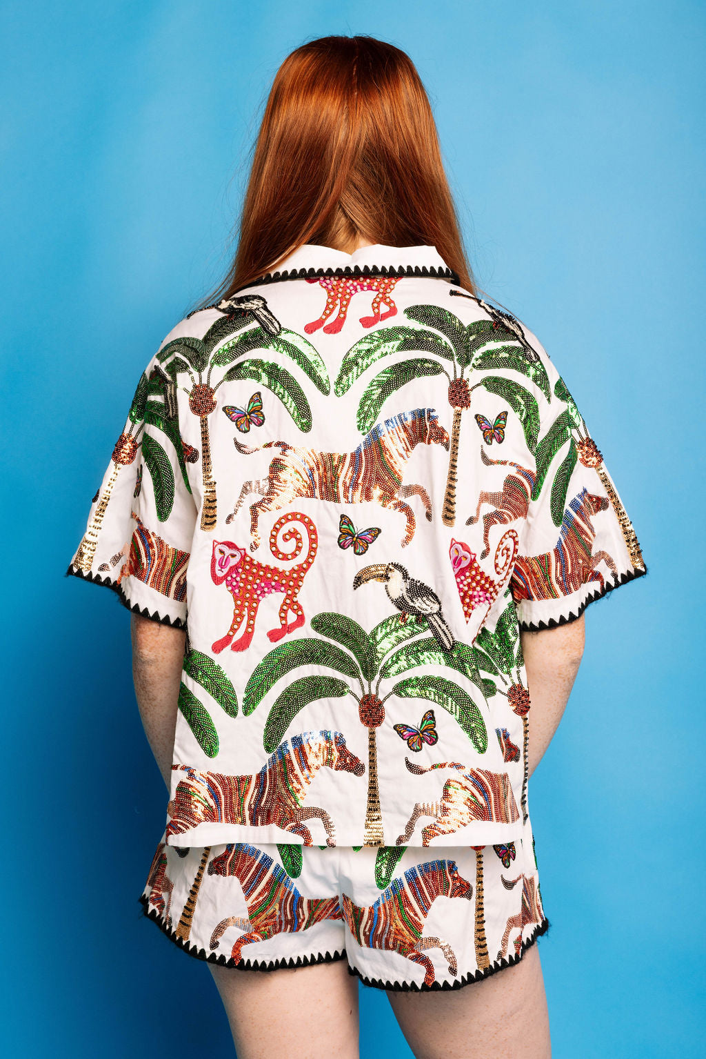 Back view of the Queen of Sparkles white safari-print button-up top and matching shorts on a blue background, showing green palm trees, colorful animal and butterfly motifs, and black trim.