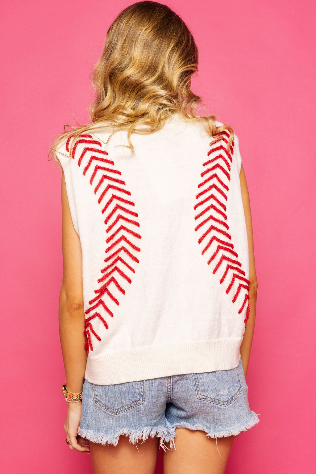 Back view of a model wearing the Queen of Sparkles white sleeveless sweater vest with red baseball-stitch detailing against a pink background.