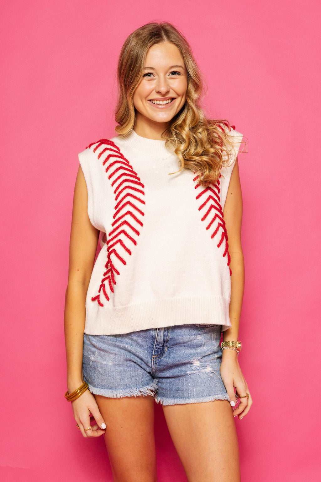 Front view of a model wearing the Queen of Sparkles white sleeveless sweater vest with red baseball-stitch detailing, styled with denim shorts against a pink background.
