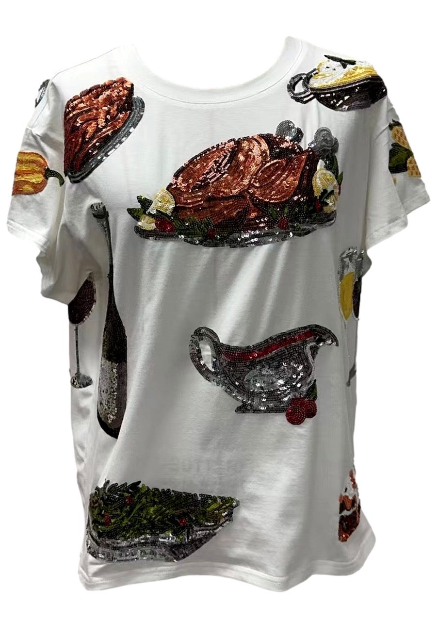 Flat-lay view of the Queen of Sparkles white Queen of Turkey tee, showing the colorful Thanksgiving meal motifs and decorative embellishments across the front.
