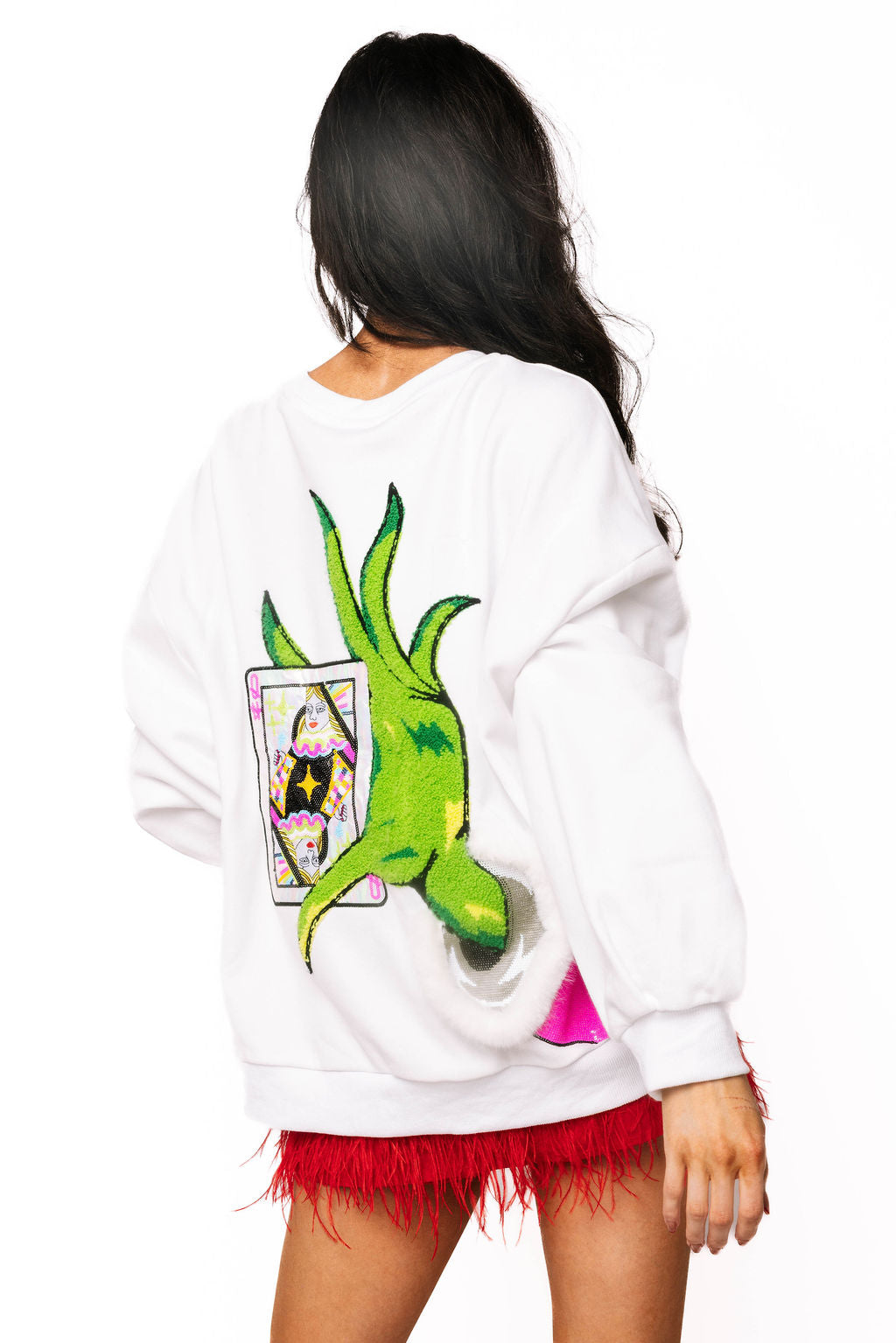 Back view of model wearing a white Christmas sweatshirt with sparklnig green Grinch hand holding a queen playing card and a pink sleeve with fluffy white trim.