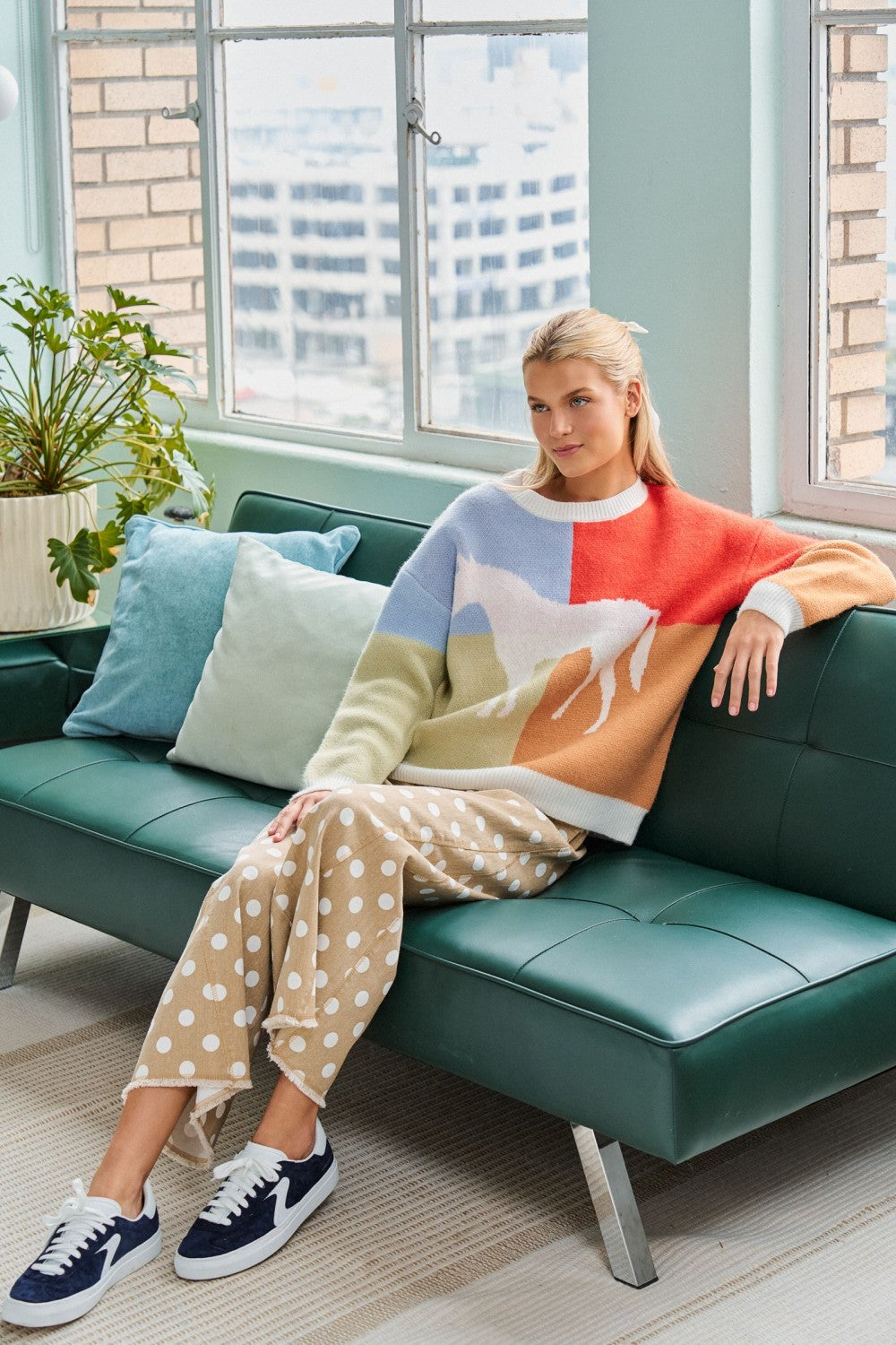 Model seated on a green sofa wearing the colorblock horse sweater with tan polka-dot pants